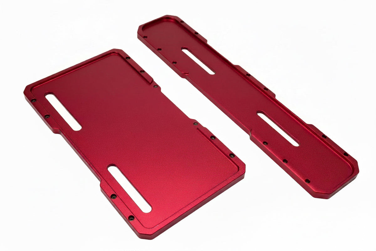 Two red anodized metal protective plates or covers with pre-drilled screw holes and cutouts, displayed on a textured bubble wrap background.
