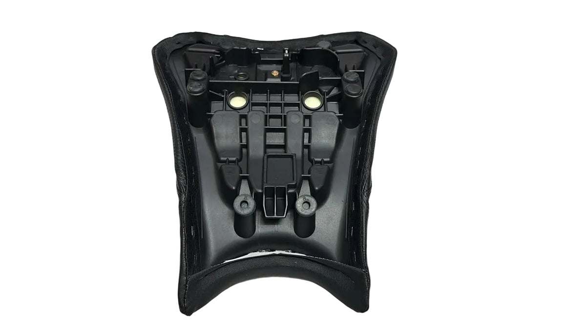 A black molded plastic motorcycle or scooter seat shell with mounting holes, shown from the underside on a white sheet, with a hand visible at the bottom left and cardboard boxes on the right.