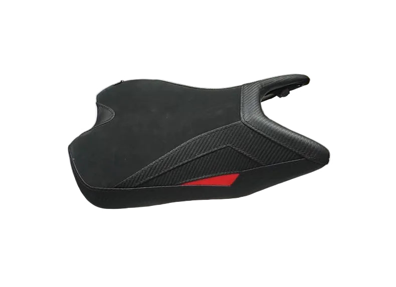 A black motorcycle or scooter seat with a textured carbon-fiber pattern and a small red accent, resting on a white plastic sheet in a warehouse setting with people partially visible.