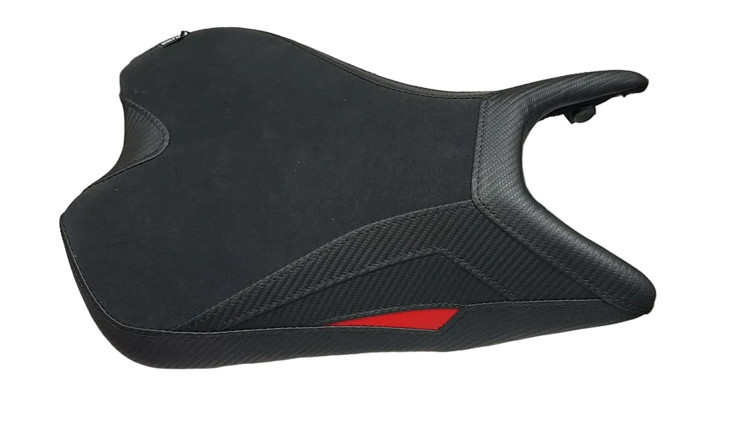A black motorcycle or scooter seat cover with a carbon fiber pattern and a small red accent, resting on a white sheet in a warehouse setting.