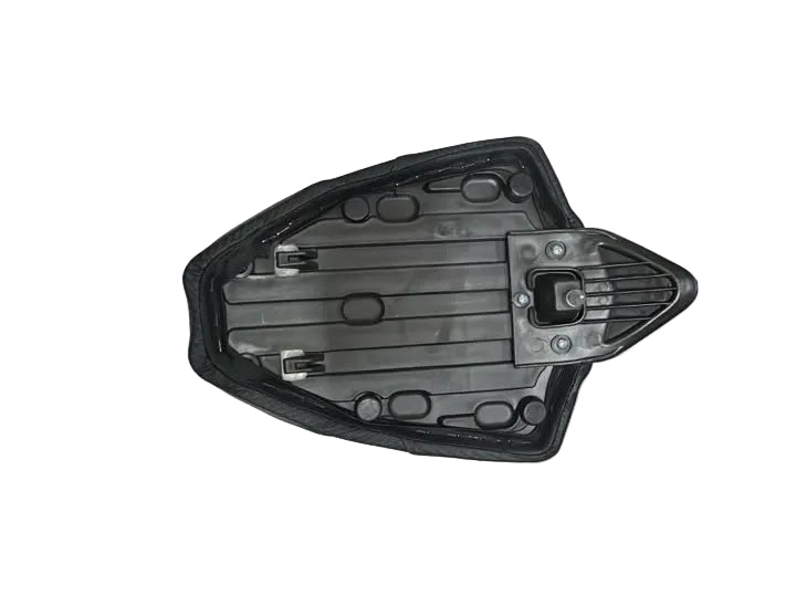 A black plastic automotive part, likely a hood ornament or trim piece, resting on a white padded surface inside a warehouse or storage area with cardboard boxes and people's legs visible.