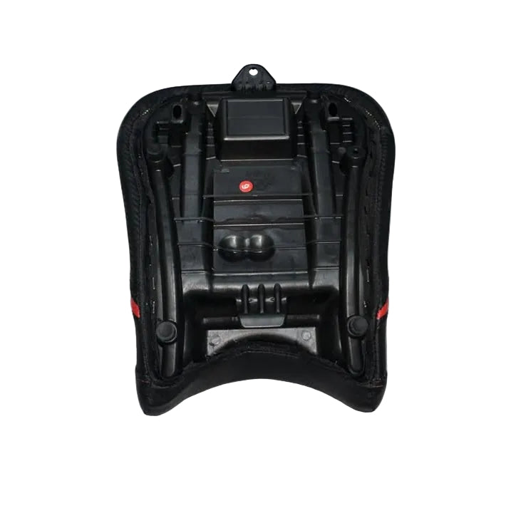 A black molded plastic seat shell or backrest component with small red accents, photographed on top of a white padded cushion in a warehouse-like setting with cardboard boxes visible below.