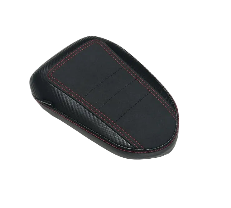 A black car center console armrest cover with red contrast stitching and carbon fiber texture details, resting on a white plastic-covered surface.