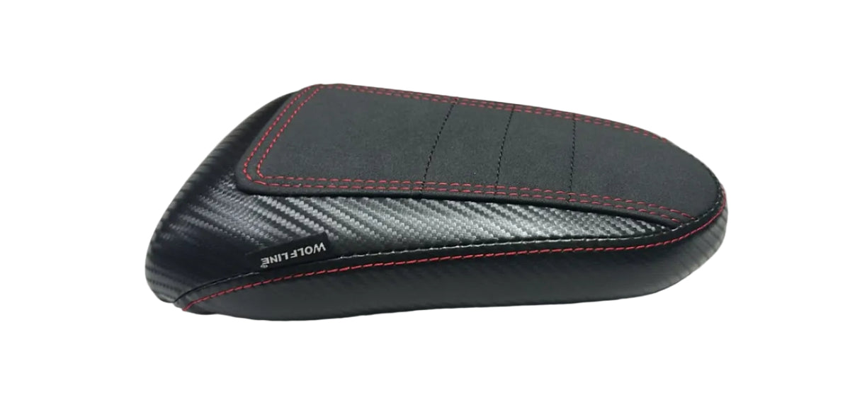 A motorcycle or scooter seat cushion cover with a black carbon-fiber pattern and red stitching, resting on a white fabric-covered surface with cardboard boxes in the background.